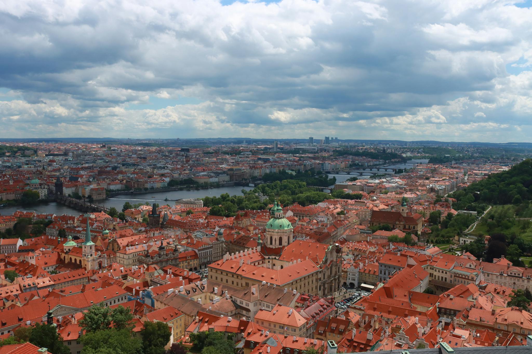Panoramic aerial view of Prague with iconic red rooftops and Vltava river under a cloudy sky.